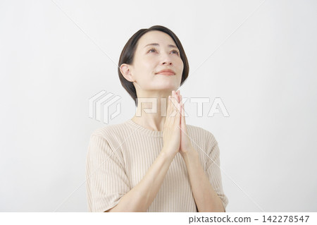 A woman in her 40s with her eyes closed and hands clasped in prayer against a white background. A woman in her 40s with her eyes closed and hands clasped in prayer against a white background. 142278547