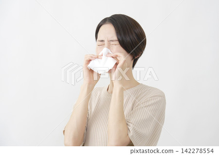 A woman in her 40s blowing her nose with a tissue against a white background. A woman in her 40s blowing her nose with a tissue against a white background. 142278554