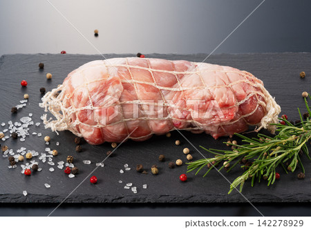 Raw pork block in netting placed on a slate plate, garnished with rosemary and spices, against a black background. Raw pork block in netting placed on a slate plate, garnished with rosemary and spices, against a black background. 142278929