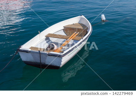 A small white rowboat moored to a buoy and floating on calm blue harbor water A small white rowboat moored to a buoy and floating on calm blue harbor water 142279469