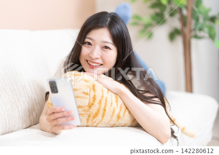 一位年輕女子躺在沙發上,面帶微笑,手裡拿著她的智慧型手機。 一位年輕女子躺在沙發上,面帶微笑,手裡拿著她的智慧型手機。 142280612