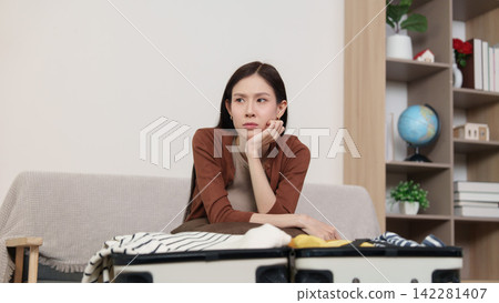 Thoughtful Asian woman sitting with open suitcase planning clothes for travel trip Thoughtful Asian woman sitting with open suitcase planning clothes for travel trip 142281407
