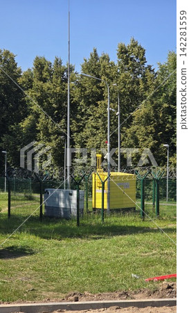 A yellow industrial electrical transformer station is enclosed behind a metal security fence in a park area. A yellow industrial electrical transformer station is enclosed behind a metal security fence in a park area. 142281559