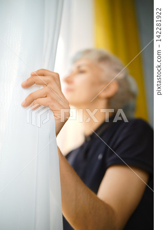 Person pulls back curtain to look outside during morning light in indoor space Person pulls back curtain to look outside during morning light in indoor space 142281922
