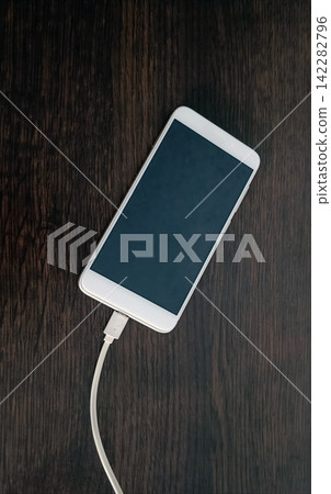 A white smartphone with a dark screen lying diagonally on a dark brown wooden surface with visible grain, connected to a white charging cable trailing downward from the device. A white smartphone with a dark screen lying diagonally on a dark brown wooden surface with visible grain, connected to a white charging cable trailing downward from the device. 142282796