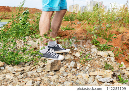 Man walking across a rocky urban construction site with weeds and exposed soil. Outdoor exploration, land development and environmental change concept. 142285109
