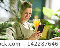 Woman Enjoying Fresh Juice and Phone in Sunlit Room 142287646