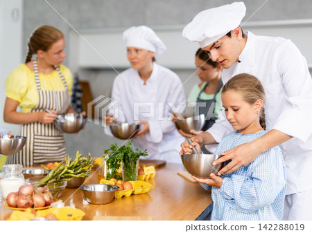 While children cooking class, male chef help small girl thoroughly stir homogeneous dough with whisk While children cooking class, male chef help small girl thoroughly stir homogeneous dough with whisk 142288019