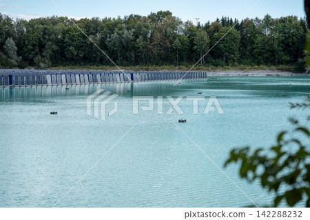 Vertical floating solar panels on the Gilching quarry lake in Bavaria, Germany, beside a gravel and crushing plant, renewable energy infrastructure, industrial landscape and innovative clean Vertical floating solar panels on the Gilching quarry lake in Bavaria, Germany, beside a gravel and crushing plant, renewable energy infrastructure, industrial landscape and innovative clean 142288232