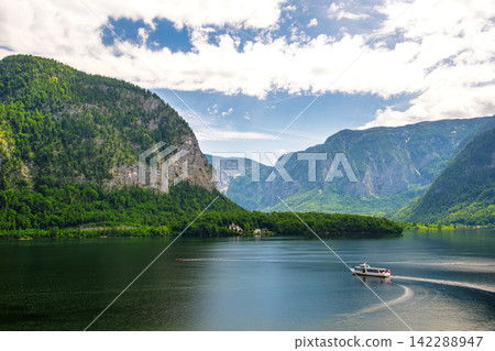 Hallstatt, Austria. A scene of a ferry sailing across Lake Hallstatt with a blue sky and lush green mountains in the background. Hallstatt, Austria. A scene of a ferry sailing across Lake Hallstatt with a blue sky and lush green mountains in the background. 142288947