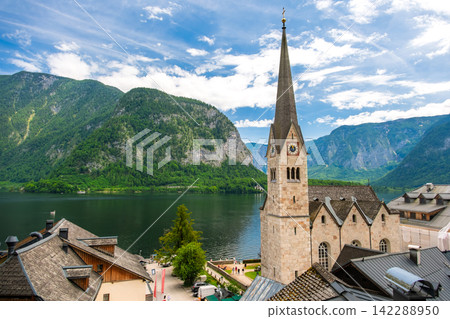 Hallstatt, Austria. The scenery of Lake Hallstatt and the village spread out against the backdrop of a blue sky and the lush green Alps. Hallstatt, Austria. The scenery of Lake Hallstatt and the village spread out against the backdrop of a blue sky and the lush green Alps. 142288950