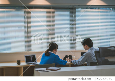 傍晚時分,辦公室裡的一個商務場景:一男一女互相展示他們的智慧型手機。 傍晚時分,辦公室裡的一個商務場景:一男一女互相展示他們的智慧型手機。 142288979