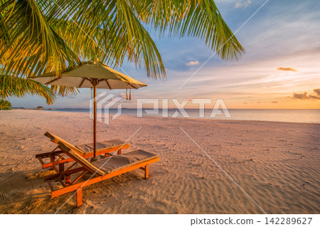 Romantic beach loungers thatched umbrellas silhouetted palms glowing golden sunset calm tropical shore. Summer honeymoon travel paradise scenic. Exotic couples getaway wall art decor Romantic beach loungers thatched umbrellas silhouetted palms glowing golden sunset calm tropical shore. Summer honeymoon travel paradise scenic. Exotic couples getaway wall art decor 142289627