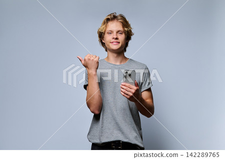 Portrait of smiling teenage student holding smartphone and pointing thumb aside Portrait of smiling teenage student holding smartphone and pointing thumb aside 142289765