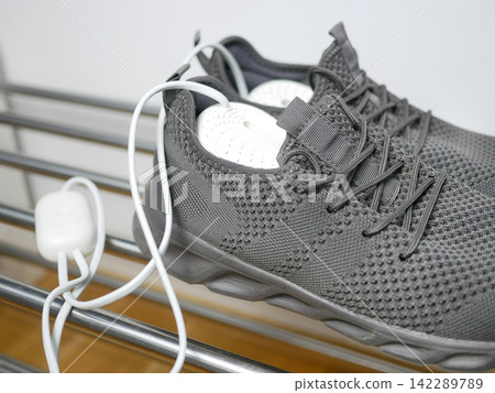 Electric shoes dryer close up. Pair of grey wet sneakers drying with a plug-in boot warmer on a shoe rack. The concept of household technologies and smart gadgets. Electric shoes dryer close up. Pair of grey wet sneakers drying with a plug-in boot warmer on a shoe rack. The concept of household technologies and smart gadgets. 142289789