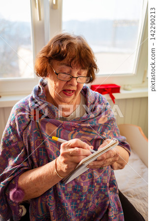 Elderly woman plans important activities with a notebook and pen at home. She continues learning and manages her personal schedule with confidence. Vertical Elderly woman plans important activities with a notebook and pen at home. She continues learning and manages her personal schedule with confidence. Vertical 142290213