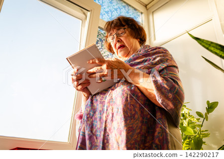 Low angle view of elderly Caucasian woman uses a notebook to manage daily responsibilities at home. Time planning, organization, and independent living concept. Low angle view of elderly Caucasian woman uses a notebook to manage daily responsibilities at home. Time planning, organization, and independent living concept. 142290217