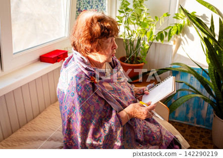 Elderly retired Caucasian woman makes handwritten notes in a notebook in a cozy living room. Productivity, learning, and managing daily tasks concept. Side view 142290219
