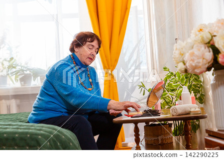 Elderly Caucasian woman enjoying personal grooming and cosmetic care while sitting on a bed. The routine emphasizes confidence, beauty and maintaining an attractive appearance in later life. Mid shot Elderly Caucasian woman enjoying personal grooming and cosmetic care while sitting on a bed. The routine emphasizes confidence, beauty and maintaining an attractive appearance in later life. Mid shot 142290225