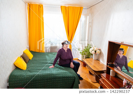 Senior Caucasian woman relaxes with virtual reality glasses after a busy day at home. Comfortable lifestyle and digital entertainment for retirees. Wide shot Senior Caucasian woman relaxes with virtual reality glasses after a busy day at home. Comfortable lifestyle and digital entertainment for retirees. Wide shot 142290237