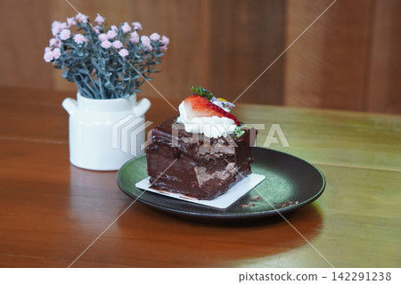 front view, chocolate cake decorated with a strawberry, and flowers and leaves, placed on a sheet of paper top a black ceramic plate, pink flowers bouquet in a ceramic cup, on brown wooden floor, front view, chocolate cake decorated with a strawberry, and flowers and leaves, placed on a sheet of paper top a black ceramic plate, pink flowers bouquet in a ceramic cup, on brown wooden floor, 142291238