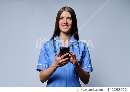 Smiling female nurse holding smartphone looking at camera against grey background 142292052