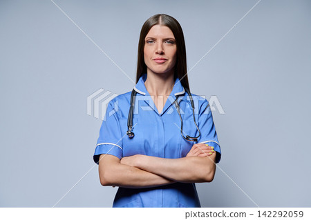 Confident female nurse in blue uniform with stethoscope standing with arms crossed against grey background Confident female nurse in blue uniform with stethoscope standing with arms crossed against grey background 142292059