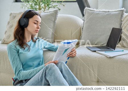 Focused teen student girl in headphones reading book sitting floor Focused teen student girl in headphones reading book sitting floor 142292111