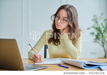 Focused female student taking notes at desk with laptop front view Focused female student taking notes at desk with laptop front view 142292128