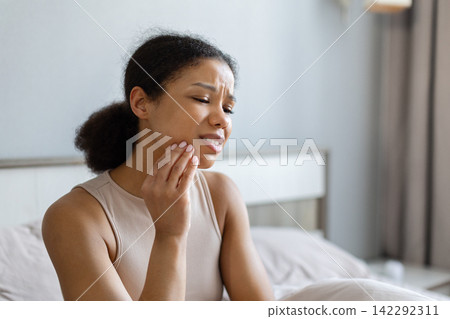 Young Black woman in bedroom touching jaw with painful expression, experiencing toothache Young Black woman in bedroom touching jaw with painful expression, experiencing toothache 142292311