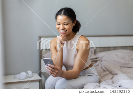 Young Black woman smiling while using smartphone in her bedroom at home Young Black woman smiling while using smartphone in her bedroom at home 142292320