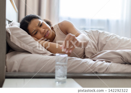 Young Black woman lying in bed reaching toward glass of water Young Black woman lying in bed reaching toward glass of water 142292322