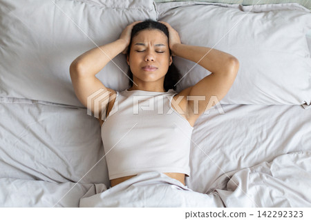 Young Black woman lies in bed holding head, experiencing headache or insomnia 142292323