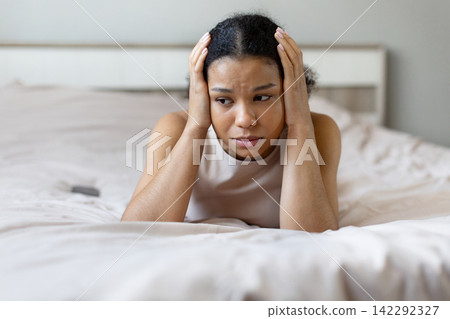 Black woman lying on bed holding head and looking worried, expressing anxiety Black woman lying on bed holding head and looking worried, expressing anxiety 142292327