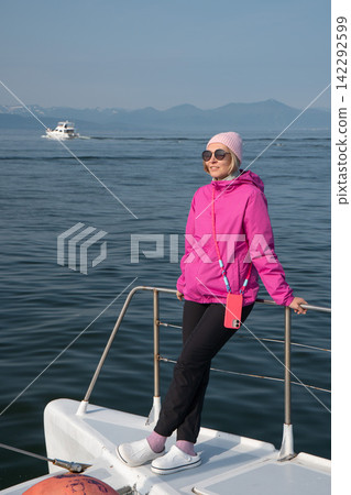 Woman in pink jacket poses on bow of catamaran with Pacific Ocean and Kamchatka mountains behind. Marine tourism, scenic travel, adventure, vacation, freedom, active outdoor lifestyle. Woman in pink jacket poses on bow of catamaran with Pacific Ocean and Kamchatka mountains behind. Marine tourism, scenic travel, adventure, vacation, freedom, active outdoor lifestyle. 142292599
