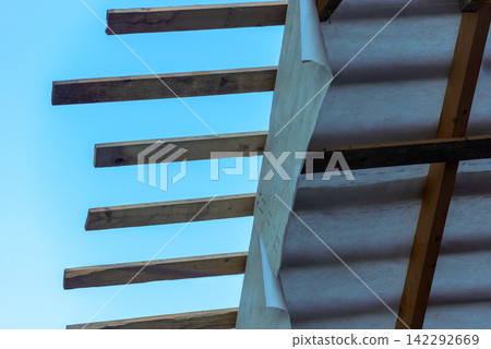 Roof structure with raw wooden rafters and white insulating material fixed to the framework, seen against a clear blue sky during a house construction or renovation project Roof structure with raw wooden rafters and white insulating material fixed to the framework, seen against a clear blue sky during a house construction or renovation project 142292669