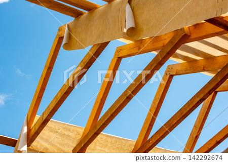 Wooden roof trusses form the structural framework of a new house, with building paper partially rolled out against a clear blue sky, symbolizing progress and growth Wooden roof trusses form the structural framework of a new house, with building paper partially rolled out against a clear blue sky, symbolizing progress and growth 142292674