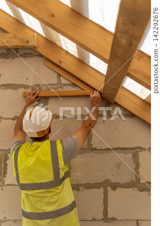 Construction worker wearing a hard hat and high visibility vest is carefully installing wooden roof rafters and beams on a new building site, focusing on structural development Construction worker wearing a hard hat and high visibility vest is carefully installing wooden roof rafters and beams on a new building site, focusing on structural development 142292676