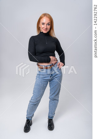 Young woman posing in a studio, wearing a black long sleeved top and light blue jeans, hands resting on her hips, expressing confidence and casual style 142292762