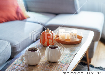 Autumn fall mood composition for hygge home decor. Hot tea or coffee drink in pumpkin's shaped cups, pumpkin decor and burning candles on the coffee table. Festive autumn, fall mood home decor Autumn fall mood composition for hygge home decor. Hot tea or coffee drink in pumpkin's shaped cups, pumpkin decor and burning candles on the coffee table. Festive autumn, fall mood home decor 142292970
