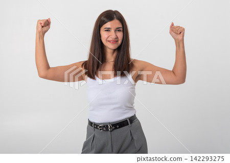 Smiling woman standing against a white background, raising her arms and flexing her biceps, conveying feelings of empowerment, determination, and success Smiling woman standing against a white background, raising her arms and flexing her biceps, conveying feelings of empowerment, determination, and success 142293275