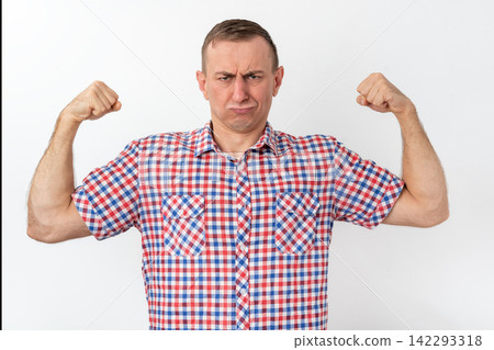 Man in a plaid shirt flexing his biceps with a humorous, pouting expression, pretending to be strong while looking mockingly weak against a clean white studio background Man in a plaid shirt flexing his biceps with a humorous, pouting expression, pretending to be strong while looking mockingly weak against a clean white studio background 142293318