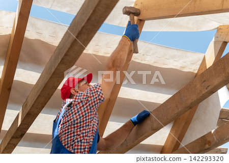 Construction worker in safety gloves and workwear precisely hitting a wooden structural beam with a hammer while building a new home or renovation project Construction worker in safety gloves and workwear precisely hitting a wooden structural beam with a hammer while building a new home or renovation project 142293320
