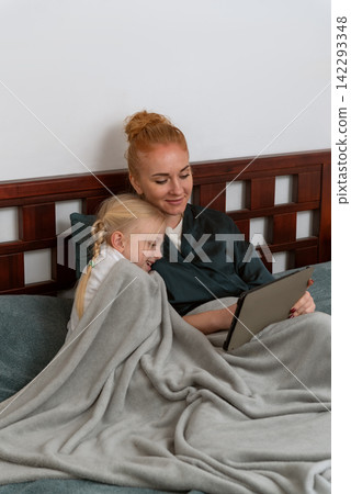 Mom and smiling daughter relaxing happily under a cozy blanket, watching a device together on the bed, creating a warm moment of family bonding and shared leisure time at home Mom and smiling daughter relaxing happily under a cozy blanket, watching a device together on the bed, creating a warm moment of family bonding and shared leisure time at home 142293348