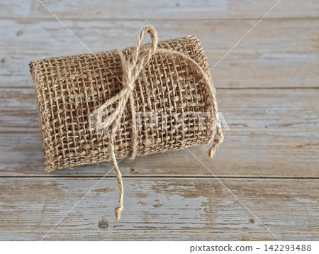 Burlap ribbon roll tied with jute twine on weathered wooden table, showing coarse woven fibers and natural textile texture for craft, gift wrapping, handmade decoration and packaging concepts. Burlap ribbon roll tied with jute twine on weathered wooden table, showing coarse woven fibers and natural textile texture for craft, gift wrapping, handmade decoration and packaging concepts. 142293488