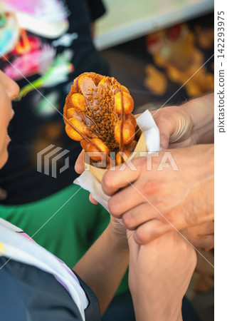 Bubble waffle dessert in hand of child. Bubble waffle dessert topped with chocolate sauce and crunchy pieces is handed to child, outdoor event Bubble waffle dessert in hand of child. Bubble waffle dessert topped with chocolate sauce and crunchy pieces is handed to child, outdoor event 142293975