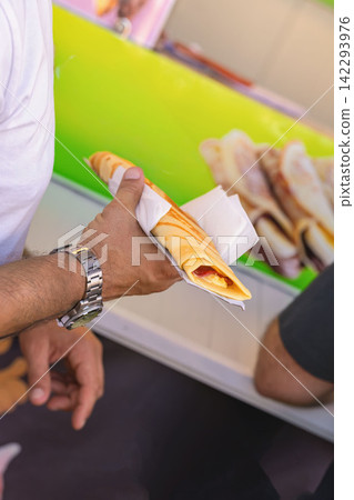 Hand of Man holding crepe. Man holding freshly prepared savory crepe wrapped in napkin, outdoor food stall. Convenient handheld street food served during summer event Hand of Man holding crepe. Man holding freshly prepared savory crepe wrapped in napkin, outdoor food stall. Convenient handheld street food served during summer event 142293976
