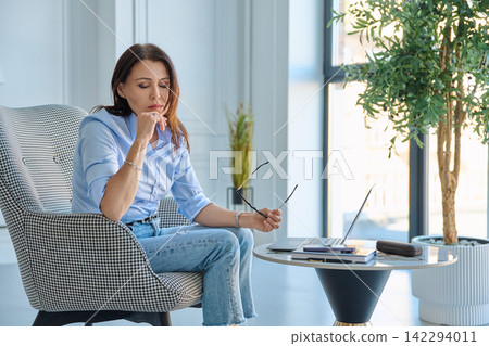 Pensive businesswoman taking off glasses feeling tired near laptop in office Pensive businesswoman taking off glasses feeling tired near laptop in office 142294011
