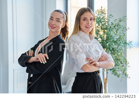 Two smiling businesswomen standing back to back with crossed arms looking at camera in modern bright office Two smiling businesswomen standing back to back with crossed arms looking at camera in modern bright office 142294223