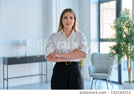 Smiling young business woman standing with arms crossed in modern light office Smiling young business woman standing with arms crossed in modern light office 142294252